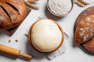 Mísa s vykynutým těstem obklopená moukou a pečicím náčiním připravená k dalšímu zpracování.