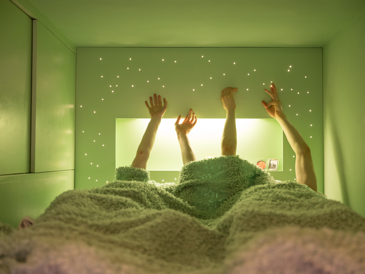 Úzký prostor ložnice s jemným zeleným osvětlením a perforovanými světelnými body na stěně.