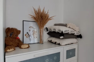 Komoda s dekorací ze sušených travin, složenými polštáři a plyšovým medvídkem v moderním interiéru.