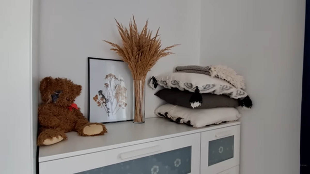 Komoda s dekorací ze sušených travin, složenými polštáři a plyšovým medvídkem v moderním interiéru.