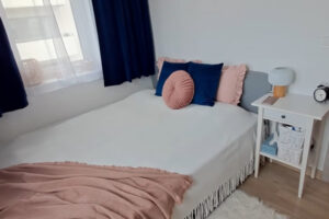 Malá ložnice s bílým povlečením, tmavě modrými závěsy a růžovými detaily.