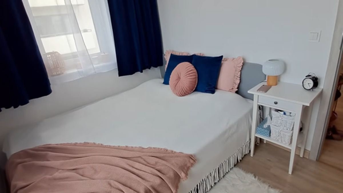 Malá ložnice s bílým povlečením, tmavě modrými závěsy a růžovými detaily.