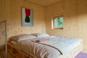 Ložnice s dřevěným obkladem, velkou postelí a malým oknem do lesa.