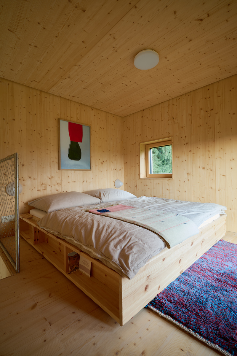 Ložnice s dřevěným obkladem, velkou postelí a malým oknem do lesa.