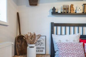 Detail nočního stolku v ložnici s proutěnou truhlou, dekoracemi ze dřeva a bílou nádobou se sušenými makovicemi.