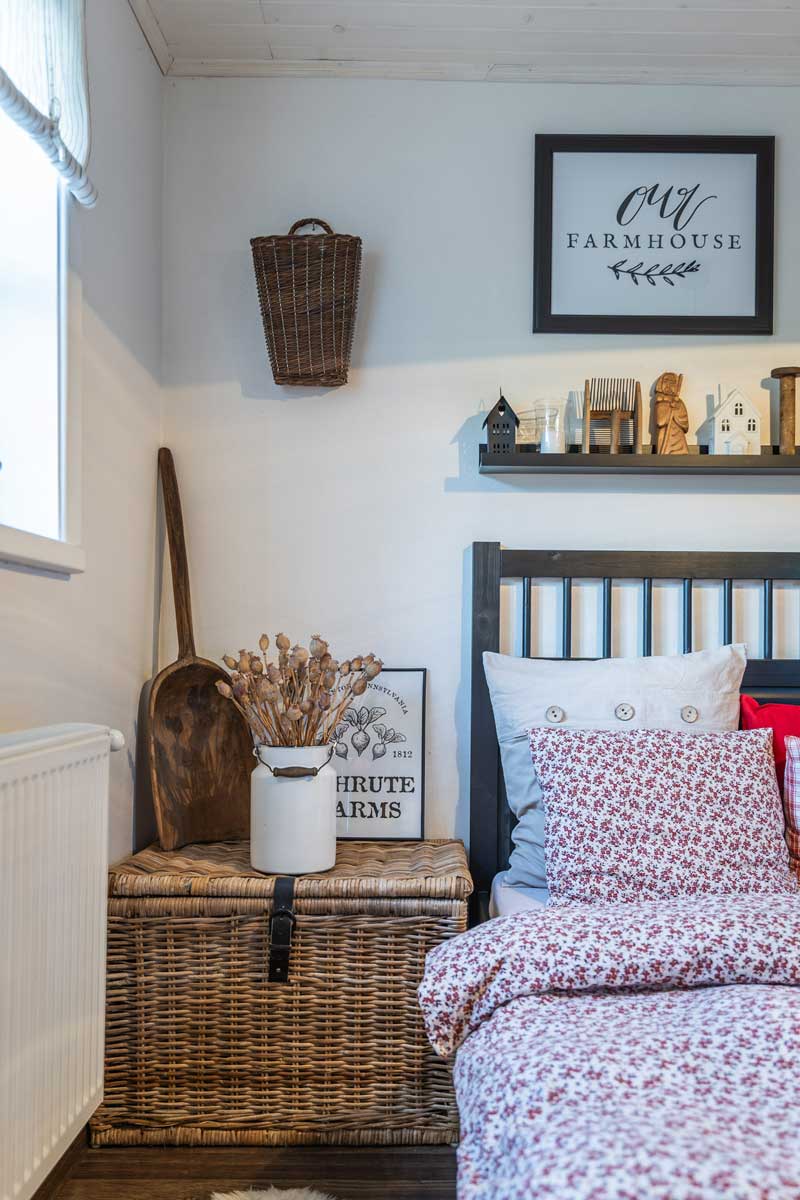 Detail nočního stolku v ložnici s proutěnou truhlou, dekoracemi ze dřeva a bílou nádobou se sušenými makovicemi.