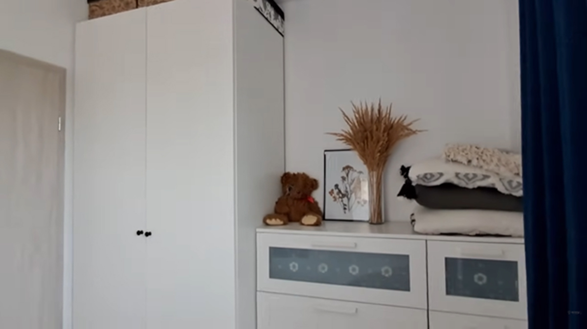 Komoda s dekorací ze sušených travin, polštáři a plyšovým medvídkem v útulné ložnici.