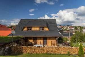 Pohled na moderní roubenku se šindelovou střechou a dřevěnou terasou v horské vesnici.