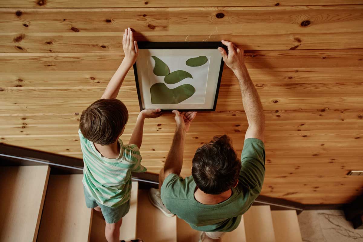 Otec se synem věší obraz na dřevěnou stěnu.