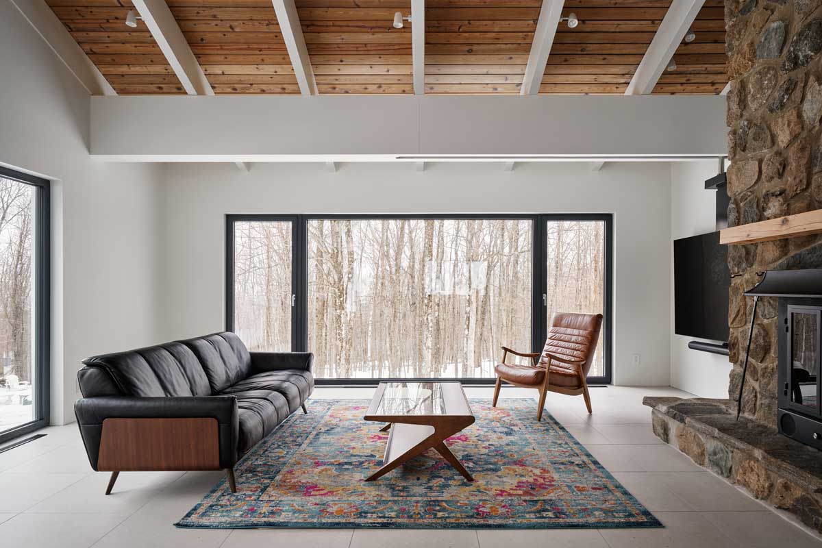 Obývací pokoj s dominantním kamenným krbem a výhledem do zasněženého lesa vytváří dokonalou kombinaci rustikálního tepla a moderní jednoduchosti.