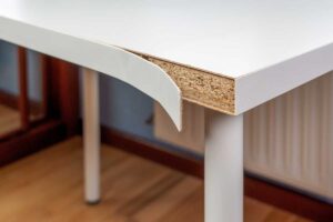 Detail odlepené bílé fólie z hrany dřevotřískové desky stolu.