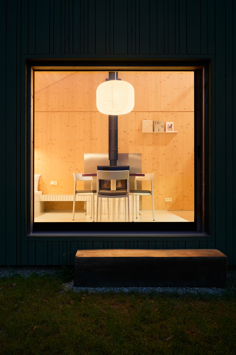 Večerní pohled do prosvětleného interiéru chaty s dřevěným obkladem a jídelním stolem.