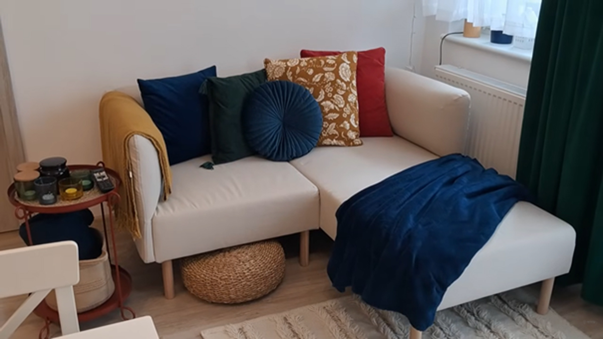 Světlá rohová pohovka s barevnými polštáři a dekou v obývacím prostoru malého bytu.