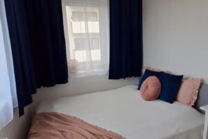 Ložnice s bílou postelí, tmavě modrými závěsy a pastelovými doplňky.