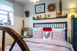 Detail čela postele s červeným květovaným povlečením a dekorativní policí plnou dřevěných figurek.