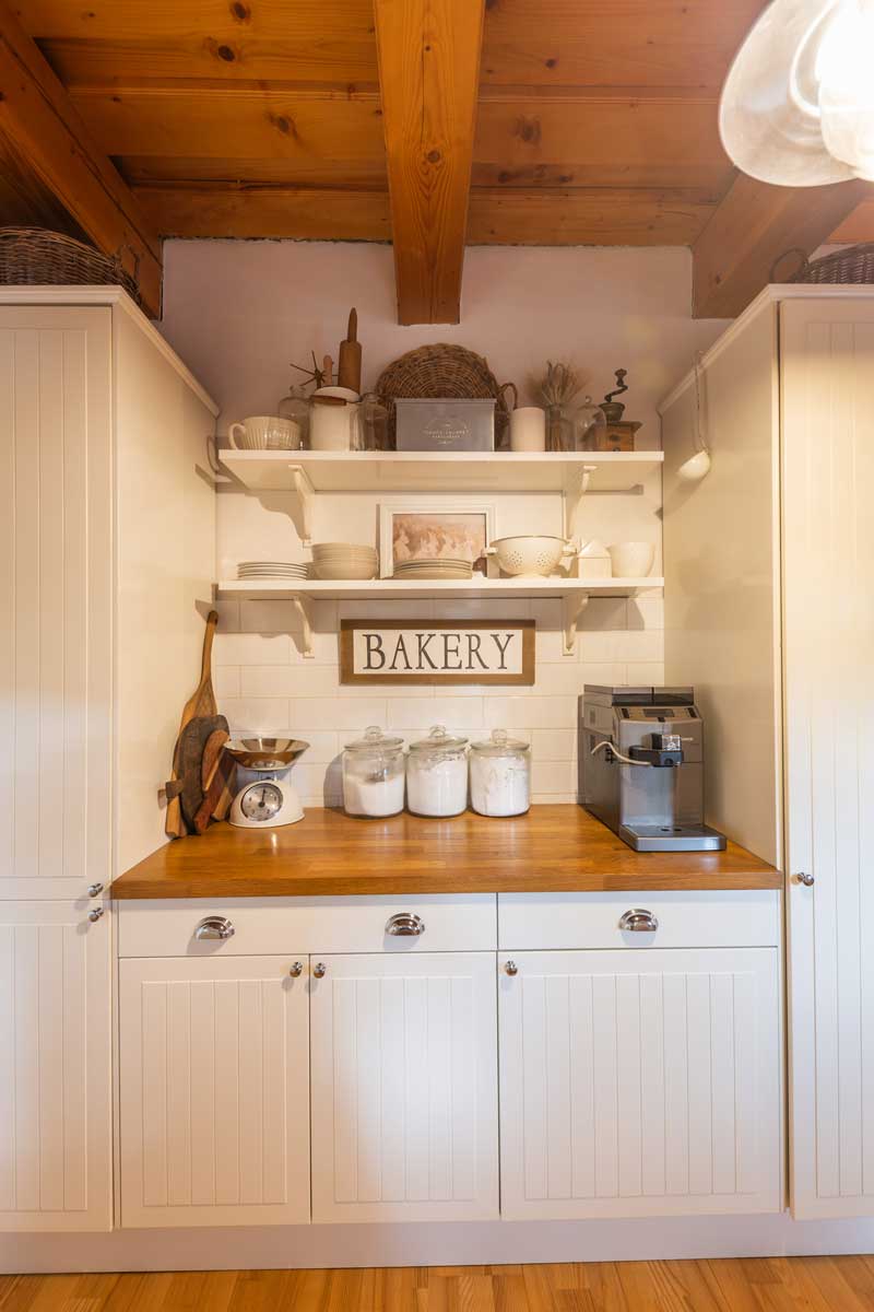 Bílá venkovská kuchyně s dřevěnou deskou, otevřenými policemi a útulnými doplňky, které odrážejí lásku majitelky k tradičnímu stylu.