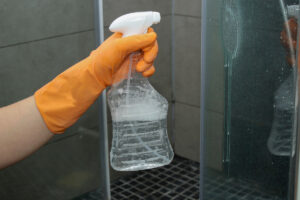 Ruka v oranžové rukavici drží rozprašovač s čisticím roztokem a nanáší ho na skleněnou stěnu sprchového koutu.