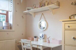 Dětský pokoj se světlým psacím stolem, jemným vzorem tapety a dekoracemi vytvářejícími klidné a příjemné prostředí.