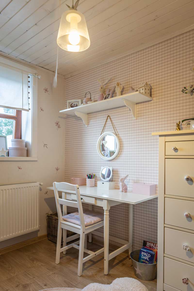 Dětský pokoj se světlým psacím stolem, jemným vzorem tapety a dekoracemi vytvářejícími klidné a příjemné prostředí.
