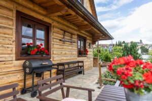 Boční terasa roubenky se dřevěným posezením a okny zdobenými červenými muškáty.