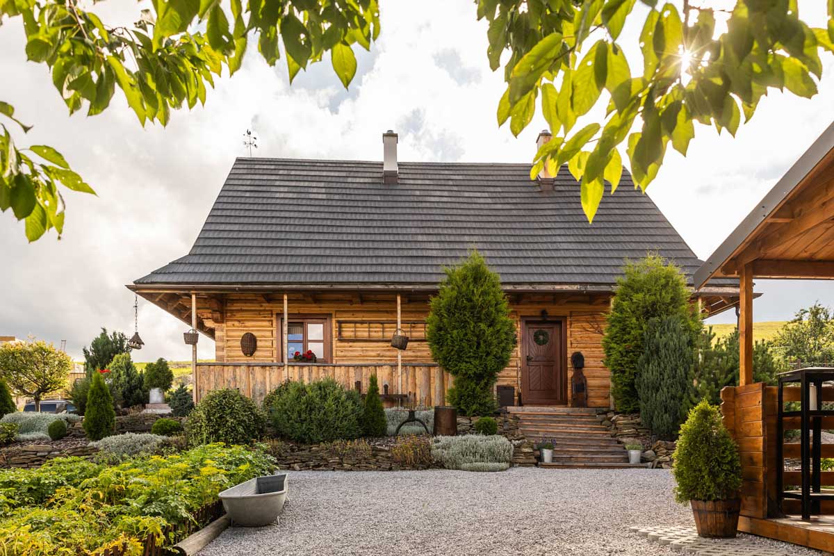 Hlavní vstup do roubeného domu obklopený zelení, dřevěnými prvky a pečlivě upravenou zahradou.