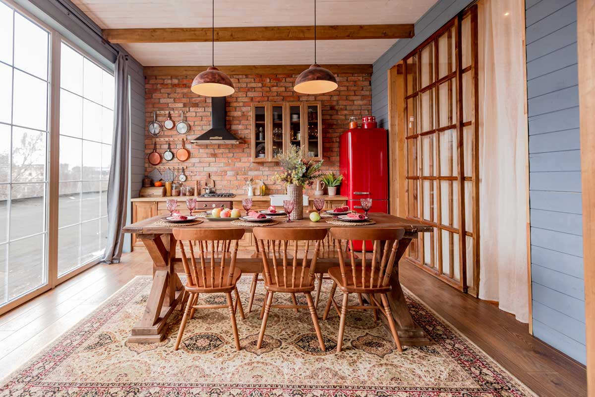Tato rustikální jídelna kombinuje masivní dřevěný stůl, cihlovou stěnu a červenou retro lednici a vytváří útulnou atmosféru cottage flow stylu.