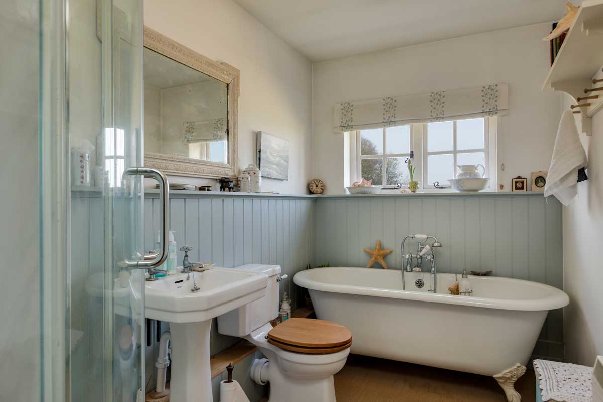 Světlá koupelna s volně stojící vanou, dřevěným obkladem a jemnými dekoracemi působí klidně a navozuje relaxační atmosféru rustikálního stylu.