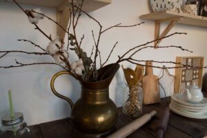 Mosazná konvice se sušenými větvičkami a dřevěnými kuchyňskými doplňky zachycuje kouzlo jednoduchých přírodních dekorací v cottage flow stylu.