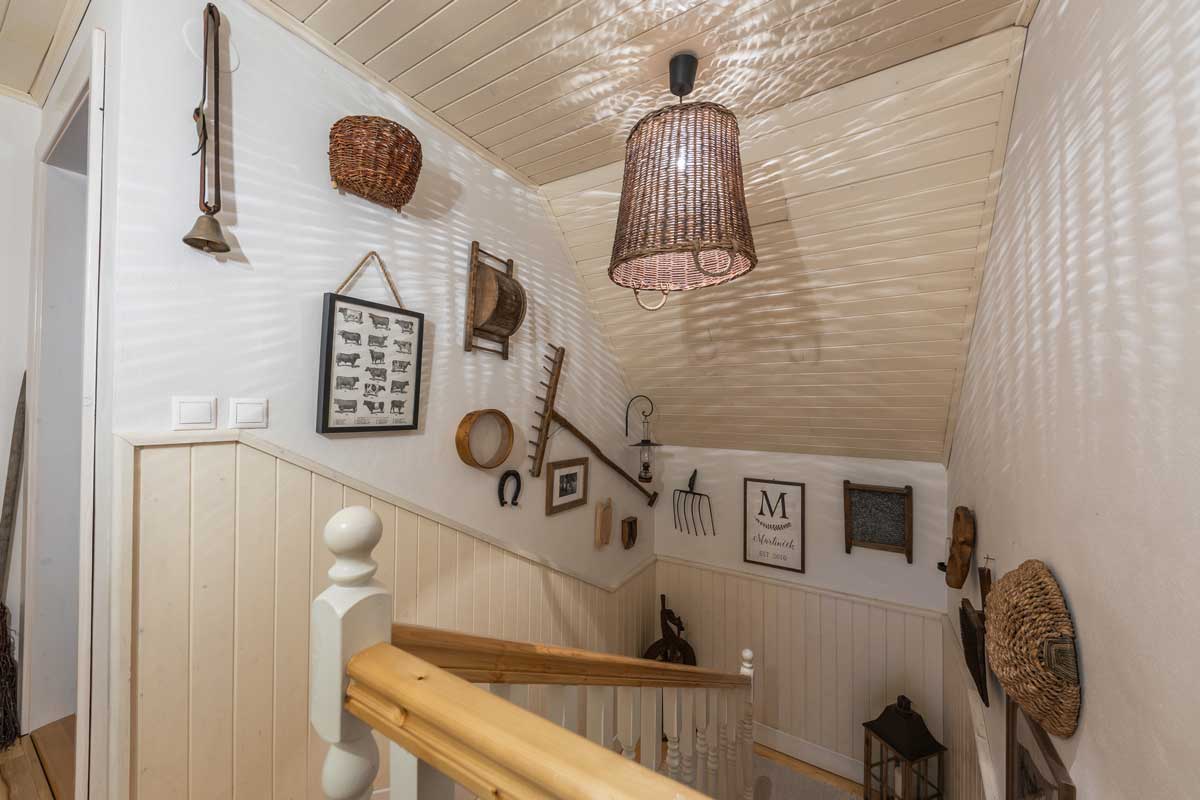 Venkovské schodiště zdobené starožitnými nástroji, košíky a proutěným stínidlem, které vytváří teplou a útulnou atmosféru rodinné roubenky.