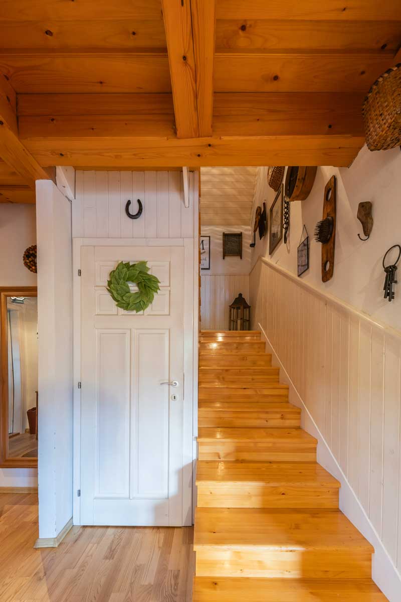 Pohled odspodu na schodiště v roubeném domě, zdobené přírodními materiály, košíky a kovovými detaily ve venkovském stylu.