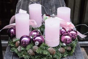 Adventní věnec s růžovými svíčkami, fialovými vánočními ozdobami a jehličím vytváří jemně barevnou variaci tradiční sváteční dekorace.