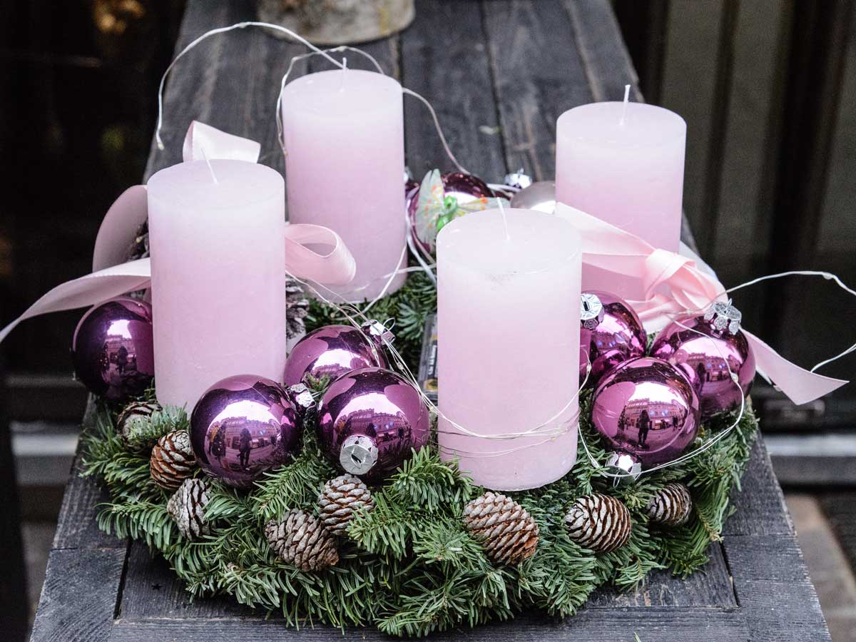 Adventní věnec s růžovými svíčkami, fialovými vánočními ozdobami a jehličím vytváří jemně barevnou variaci tradiční sváteční dekorace.