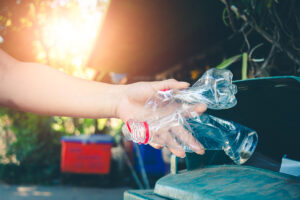 Ruka vhazující prázdné plastové lahve do kontejneru na tříděný odpad na slunci.