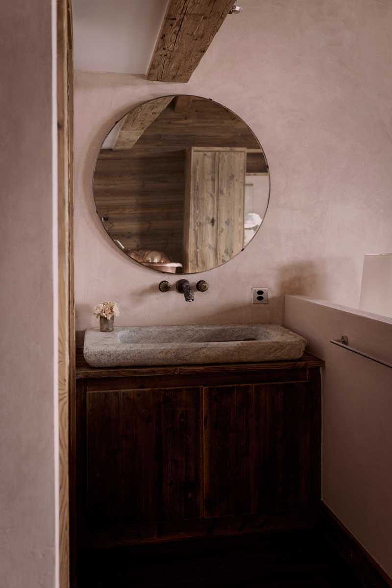 Recyklované kamenné umyvadlo a kulaté vintage zrcadlo v koupelně Chalet 1936 s přírodními povrchy.