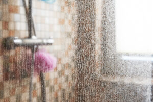 Detail skleněné stěny sprchového koutu pokryté vrstvou vodního kamene s rozostřeným pozadím koupelny.