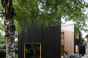 Pohled na dům s černou fasádou a velkým stromem v popředí, který tvoří součást zahrady.