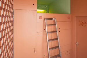 Kovový žebřík vedoucí k horní ložnicové nice v kompaktním bytě s oranžovým nábytkem.