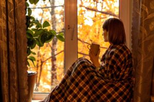 Žena sedící u okna zabalená v dece se šálkem v ruce.