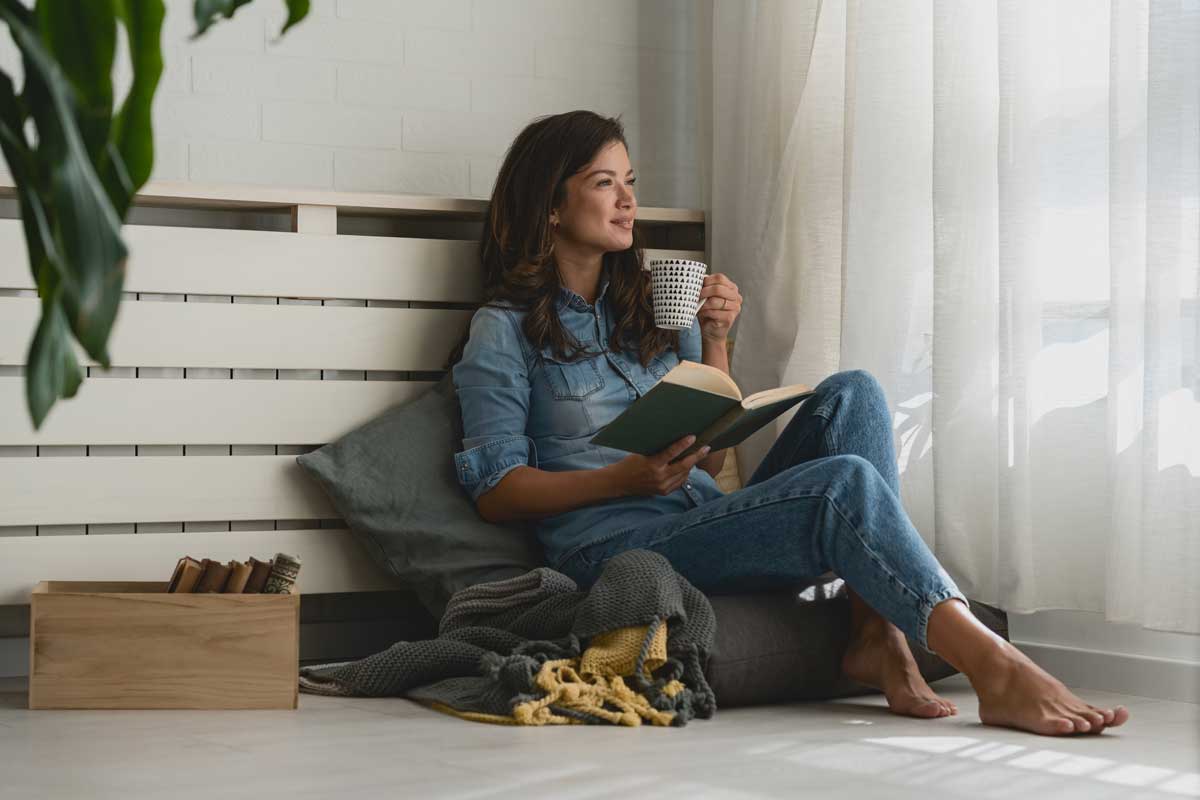 Žena sedící u topení s knihou – relaxační zimní koutek pro introverty.