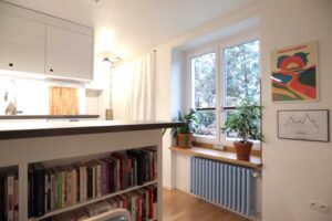 Denní zóna garsonky s okny, rostlinami, bílými závěsy a výhledem na kuchyňský prostor.