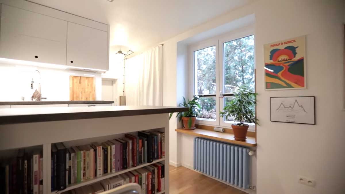 Denní zóna garsonky s okny, rostlinami, bílými závěsy a výhledem na kuchyňský prostor.