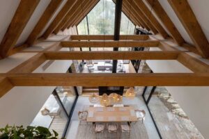 Pohled z patra na jídelní stůl, krb a velká okna v moderní horské chatě