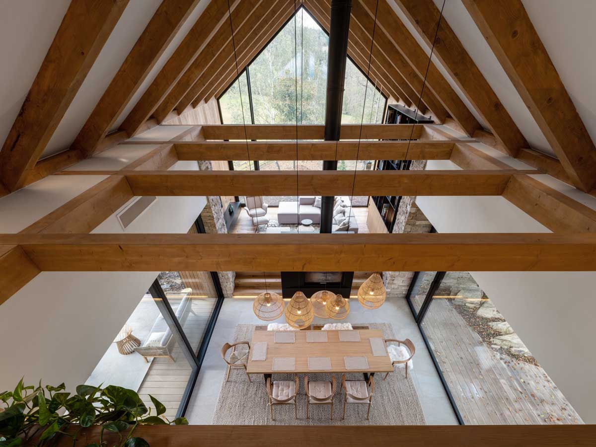 Pohled z patra na jídelní stůl, krb a velká okna v moderní horské chatě
