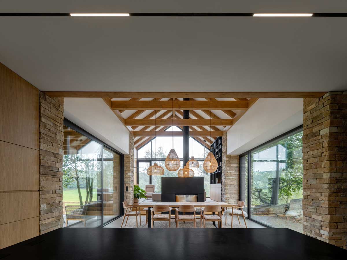 Pohled z kuchyně do jídelny s krbem a dřevěnými trámy v moderní novostavbě