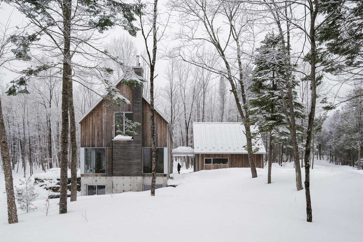 Dva objemy moderní dřevostavby kopírující svažitý terén, propojené krytou chodbou uprostřed zimní přírody.