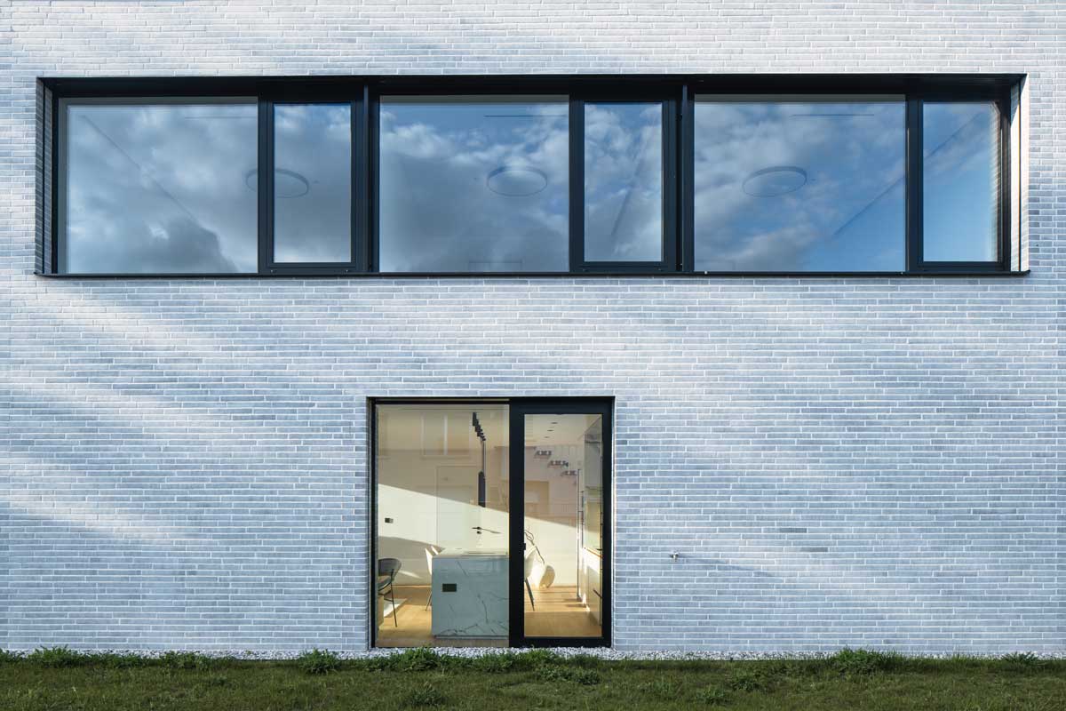 Detail fasády rodinného domu s bílou cihlovou strukturou a velkými okny v černých rámech.