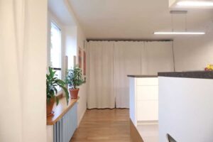 Pohled z kuchyně do hlavní obytné části garsonky s plakáty, okny a závěsem skrývajícím šatnu.