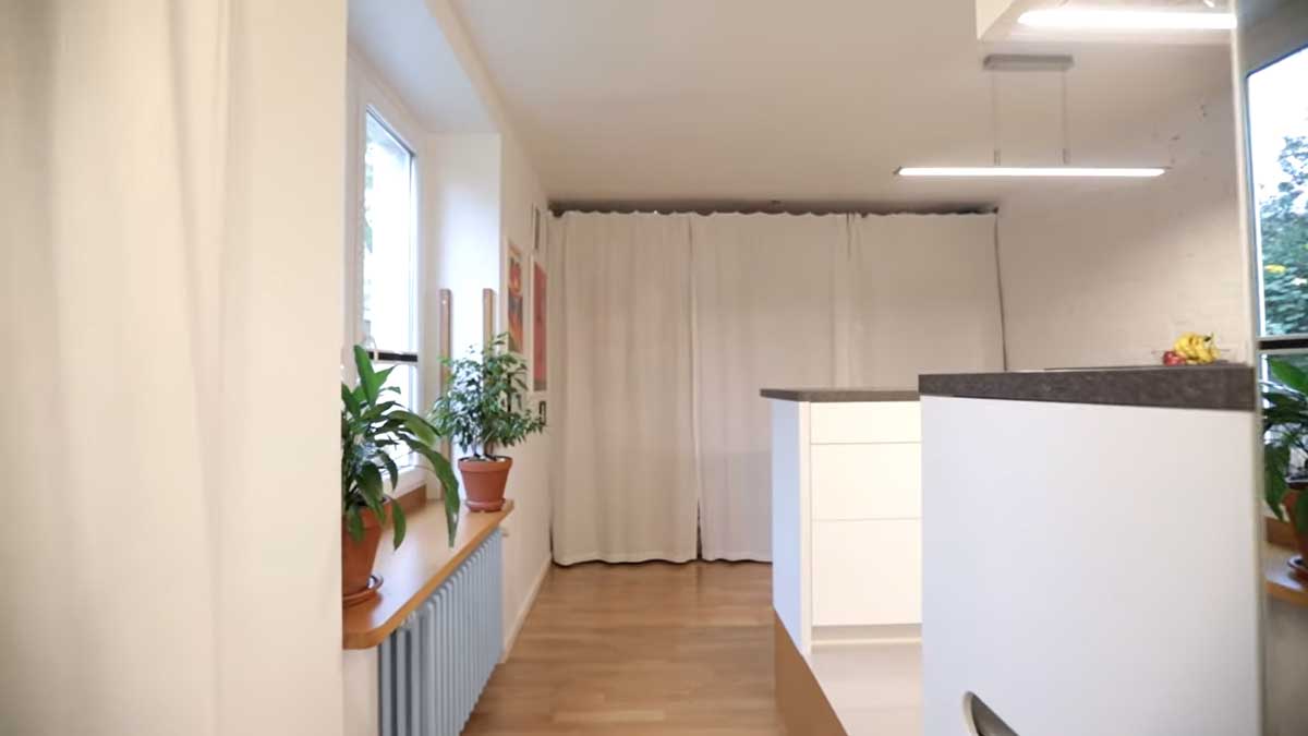 Pohled z kuchyně do hlavní obytné části garsonky s plakáty, okny a závěsem skrývajícím šatnu.