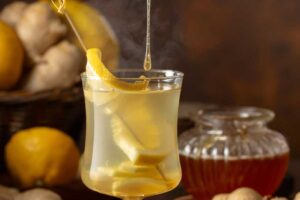 Sklenice horkého medovo-zázvorového nápoje s plátky citronu a zázvoru, do kterého stéká lžička medu.
