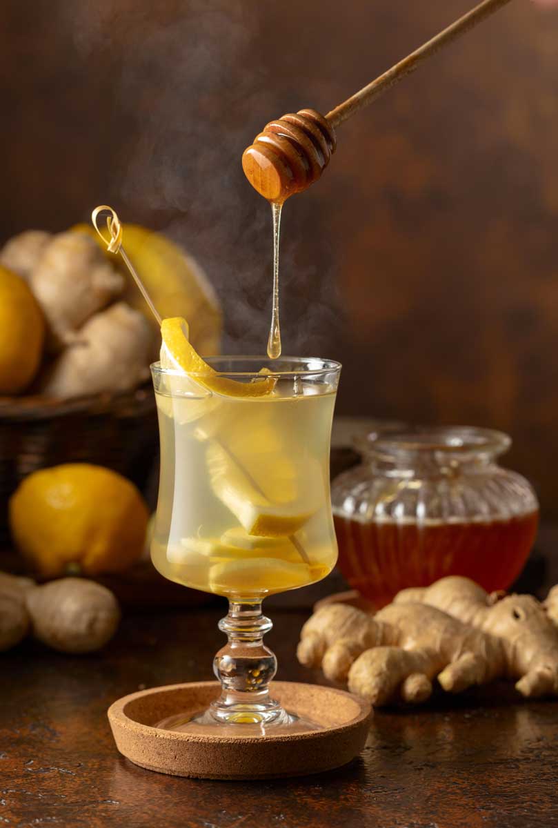 Sklenice horkého medovo-zázvorového nápoje s plátky citronu a zázvoru, do kterého stéká lžička medu.